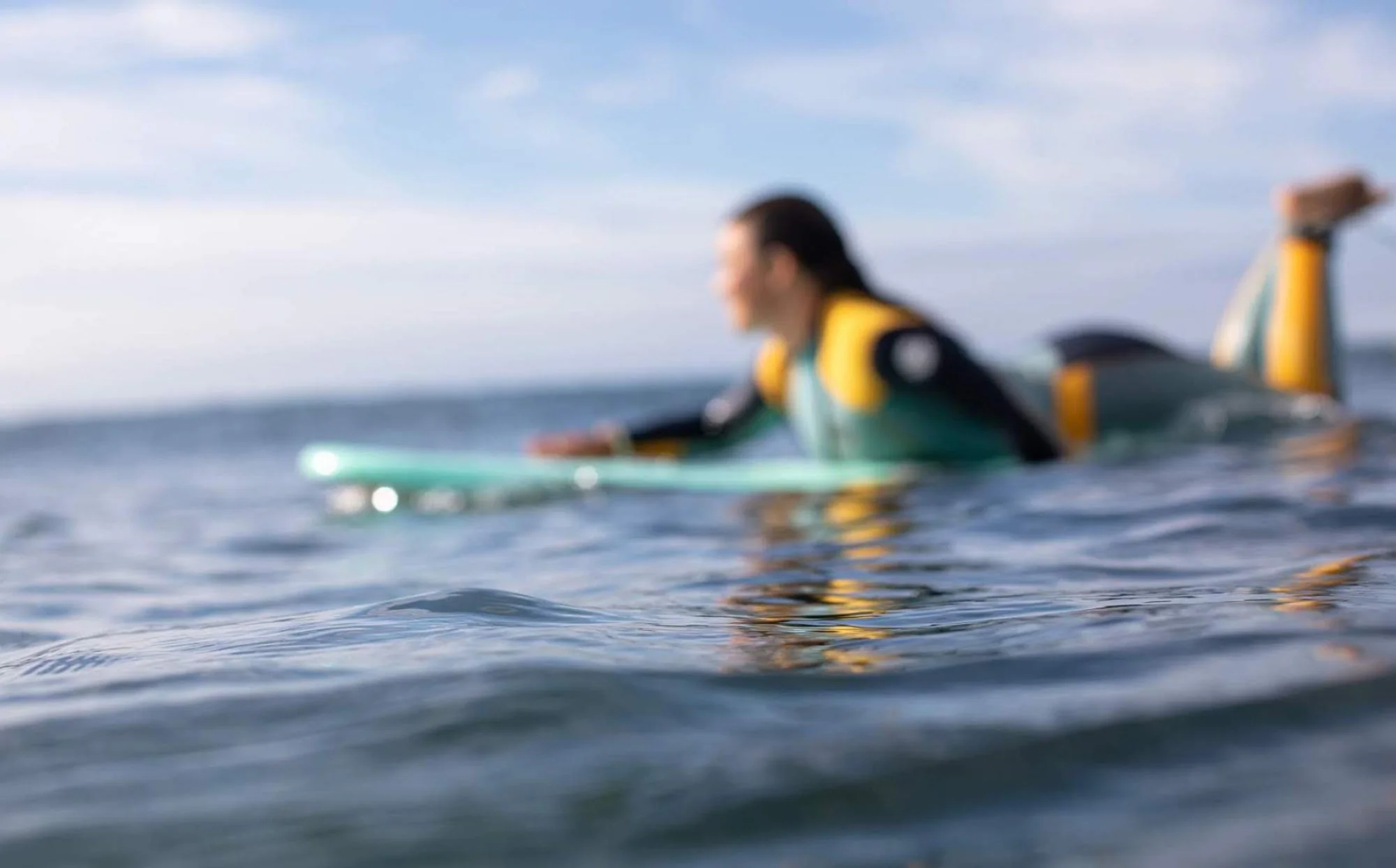 A person wearing a wetsuit lying on a surfboard in the water, with blurred ocean and sky in the background.