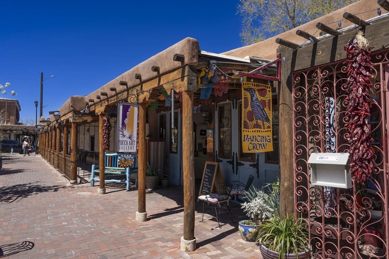 Exterior of an art gallery called Yucca Art Gallery with colorful banners, potted plants, and outdoor seating on a sunny day.