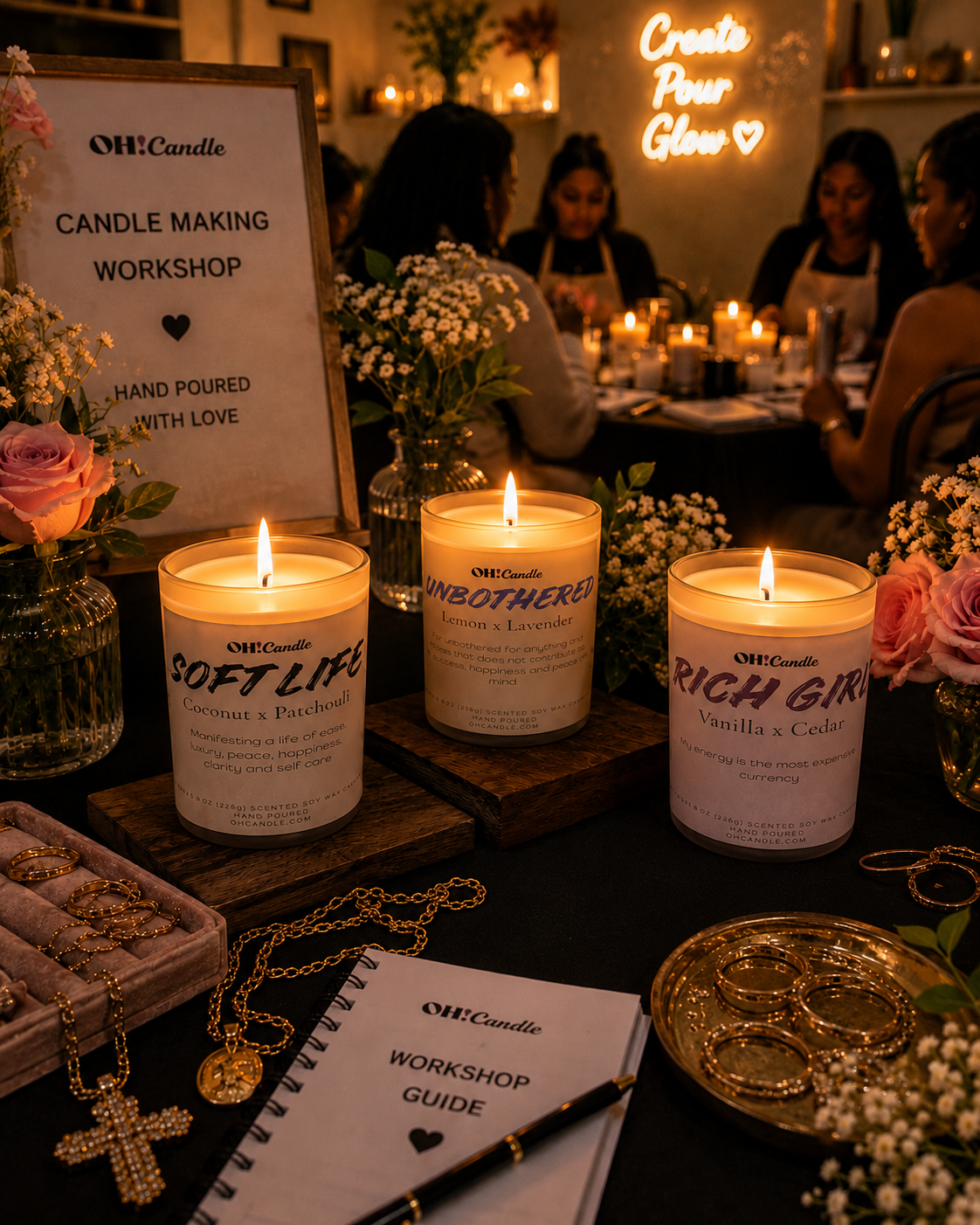 A candle-making workshop setup with three lit candles labeled 'SOFT LIFE', 'UNBOTHERED', and 'RICH GIRL'. The table is decorated with flowers, jewelry, and a workshop guide. In the background, women are seated around a table with more candles, and a neon sign on the wall reads 'Create Pour Glow'.