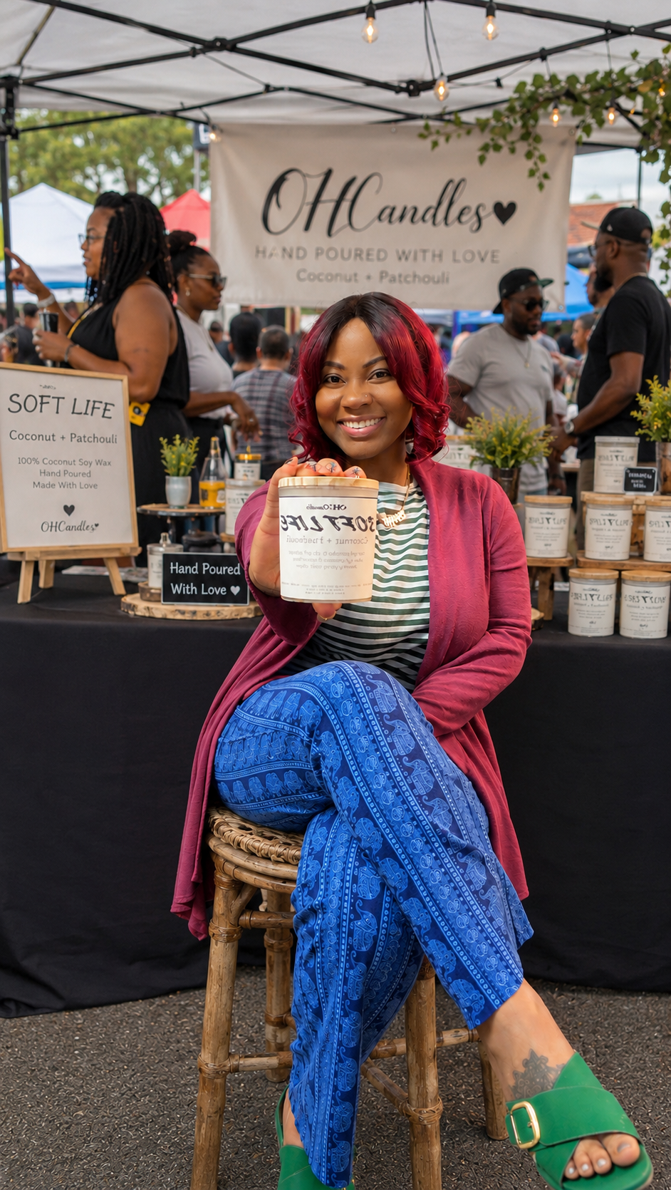 A woman with red hair sitting on a stool at an outdoor market booth for OH Candles, holding a candle jar and smiling at the camera. The booth displays candles and signs advertising hand-poured candles with coconut and patchouli scent. Other vendors and customers are visible in the background.