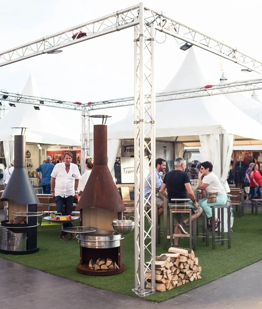 Scene of an outdoor market or festival with white tents, people sitting on high stools, and two large wood-fired outdoor grills and heaters.