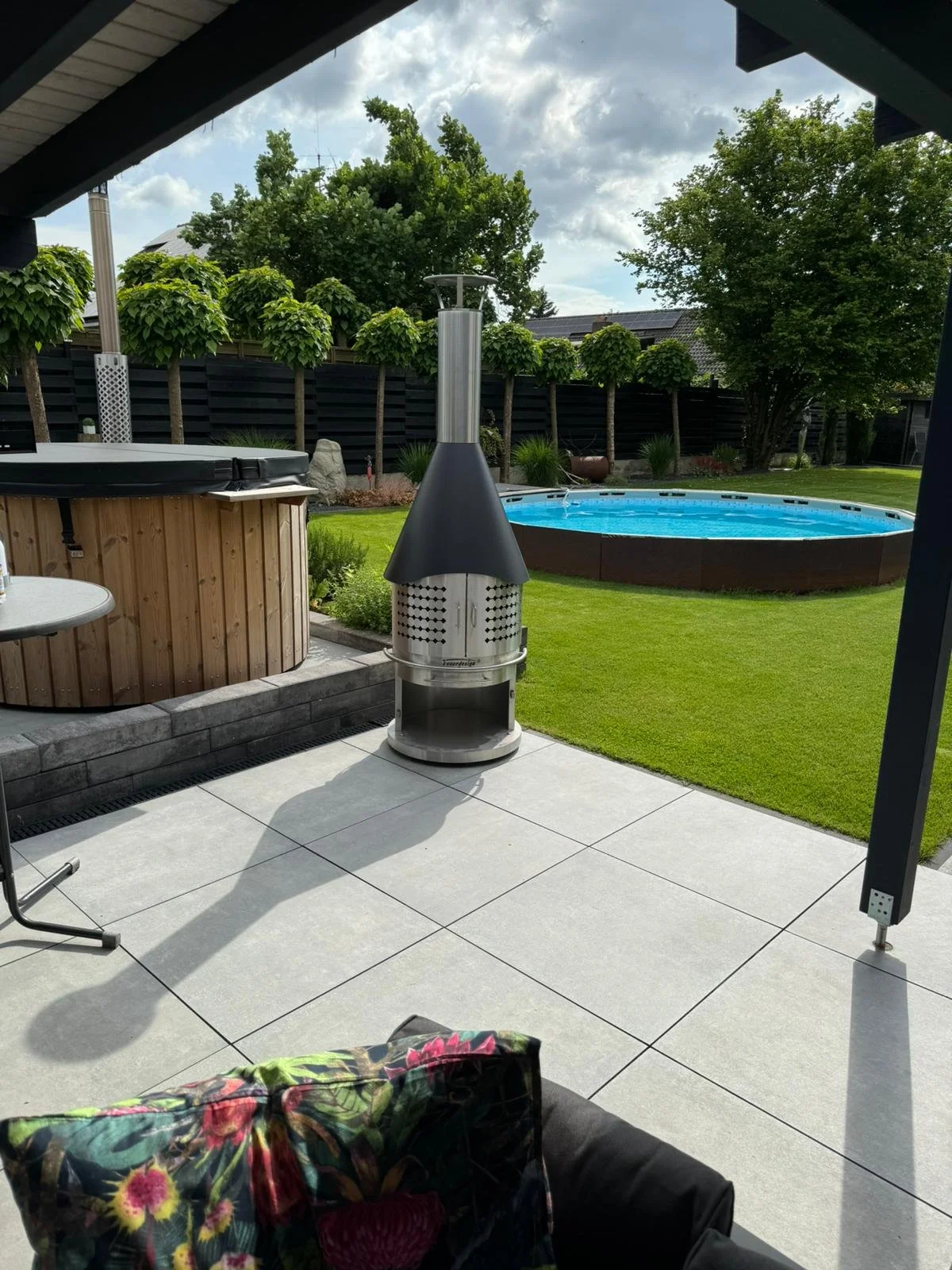 View from a patio showing a hot tub, a freestanding stainless steel outdoor heater and grill, a lawn, a round swimming pool, trees, and a cloudy sky.