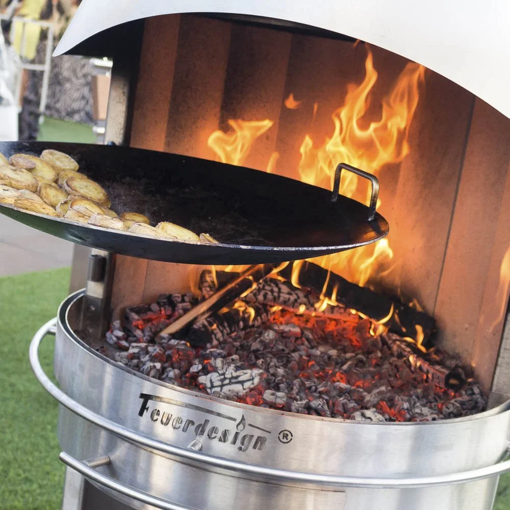 A wok with sliced potatoes cooking over an open flame in a wood-fired outdoor grill and heater.