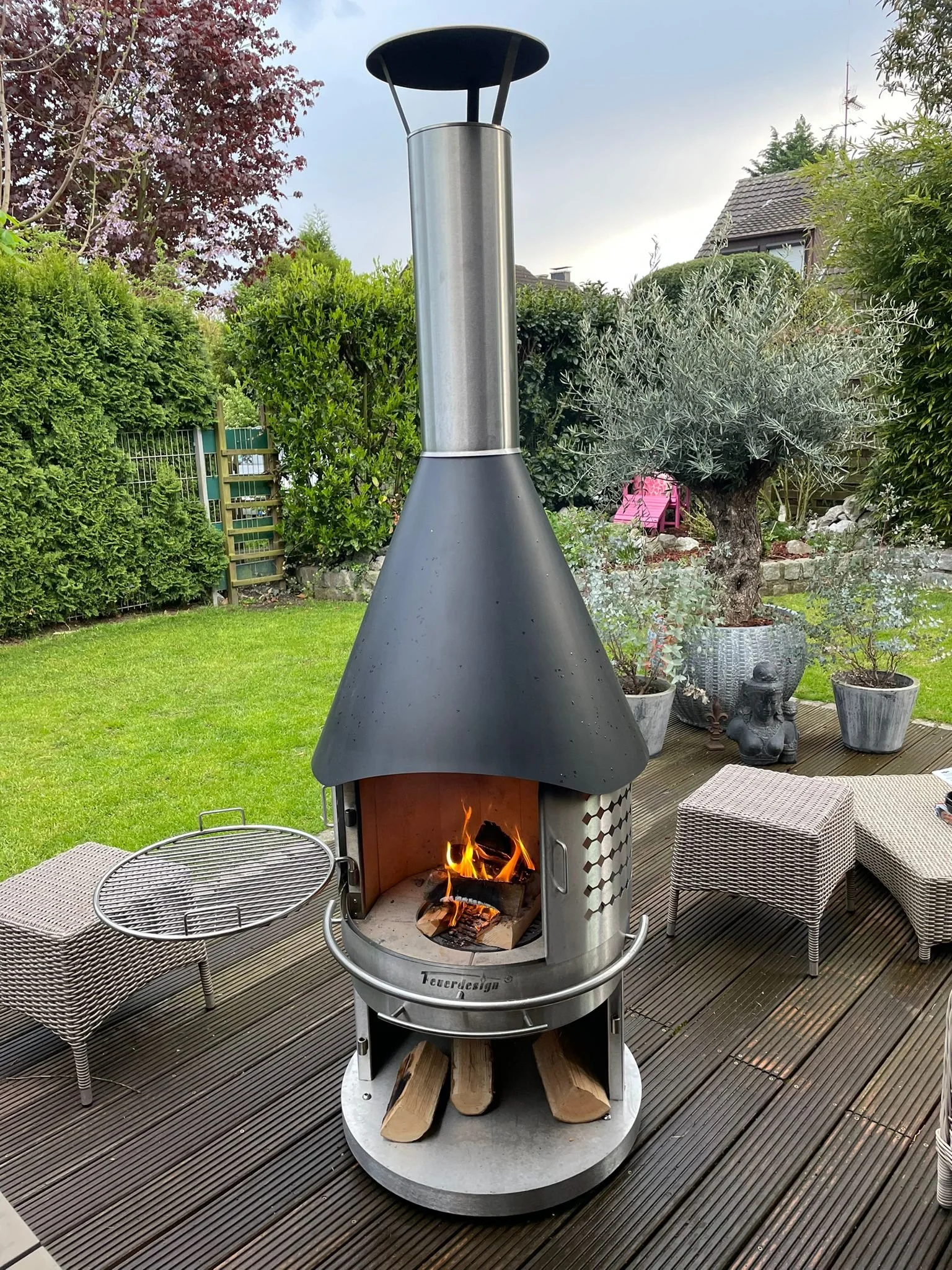 A modern outdoor fireplace with a chimney on a wooden deck, surrounded by outdoor furniture and potted plants in a garden.