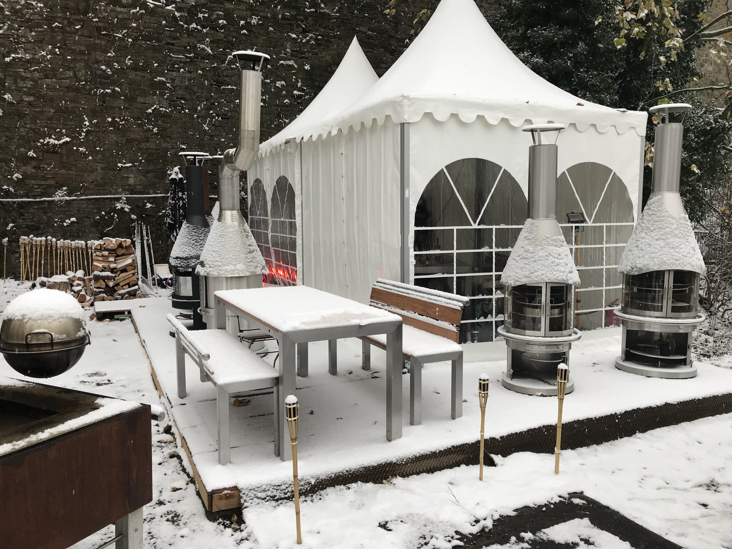 Snow-covered outdoor patio with a white gazebo, outdoor table, and chairs, outdoor furniture, surrounded by garden tools, wood logs, and outdoor heaters, with snow on the ground and trees in the background.