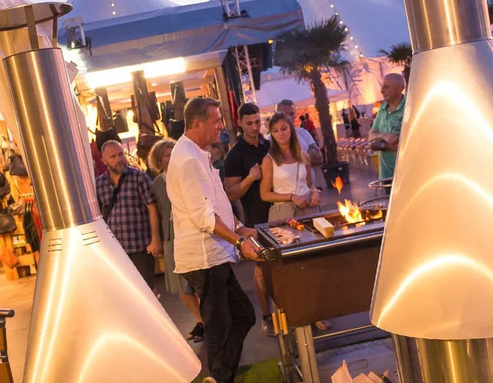 People gathered at an outdoor barbecue grill, with string lights and palm trees in the background, enjoying a social event.