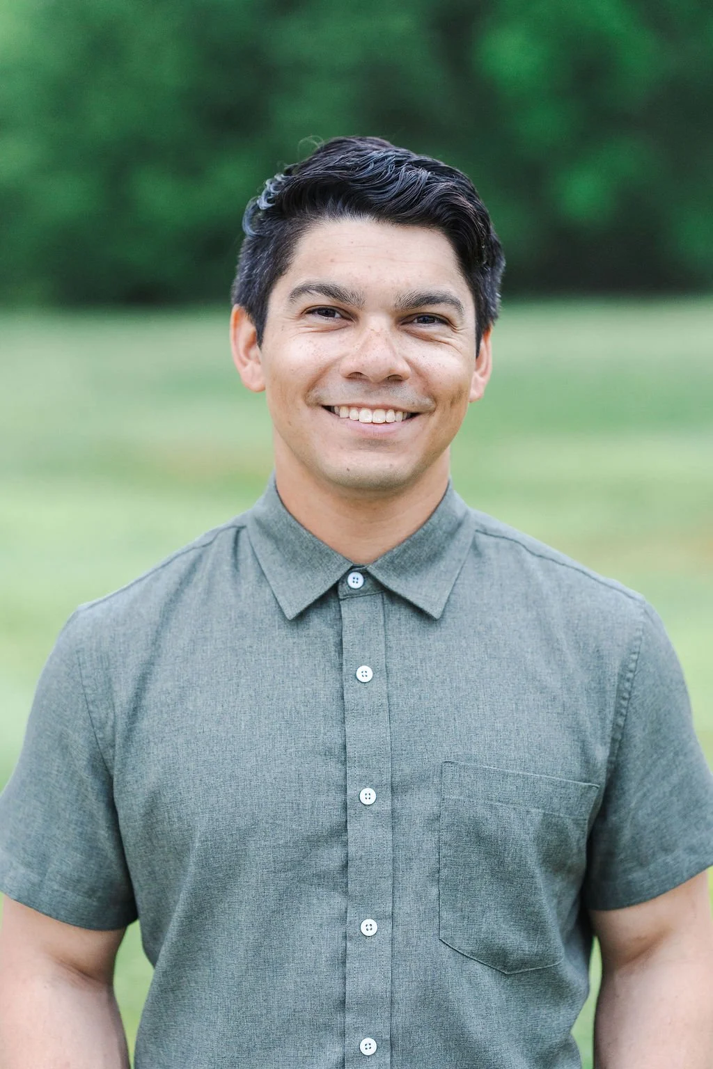 A young man with dark hair, smiling, wearing a green button-up shirt, standing outdoors with a blurred green background.