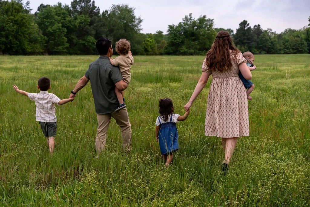 Family of six walking through a grassy field, holding hands, with trees in the background.