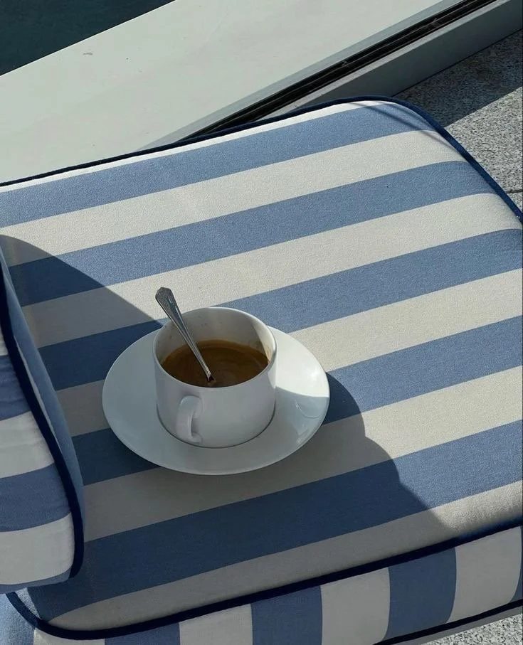 A cup of coffee with a spoon on a white saucer resting on a blue and white striped outdoor lounge cushion.