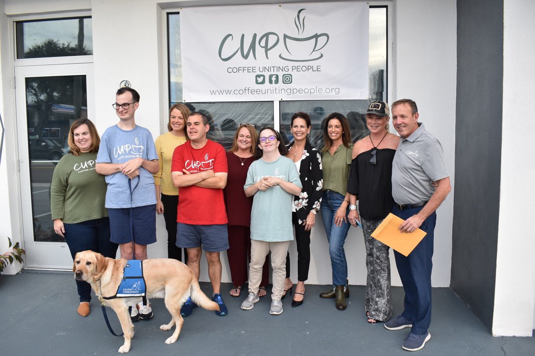 Group of nine people standing outside a building with a banner that says "Coffee Uniting People." They are smiling, and one person is holding an orange folder. A service dog wearing a blue vest is in front of the group.