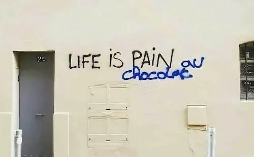 Graffiti an einer weißen Wand mit der Aufschrift "LIFE IS PAIN" und abgedecktem Wort "Chocolate" darunter.