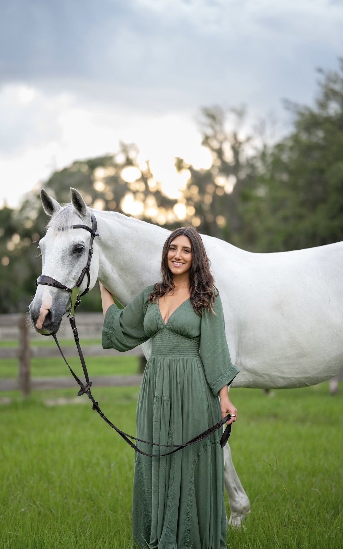 A woman in a long green dress standing beside a white horse in a grassy field with trees and a cloudy sky in the background.