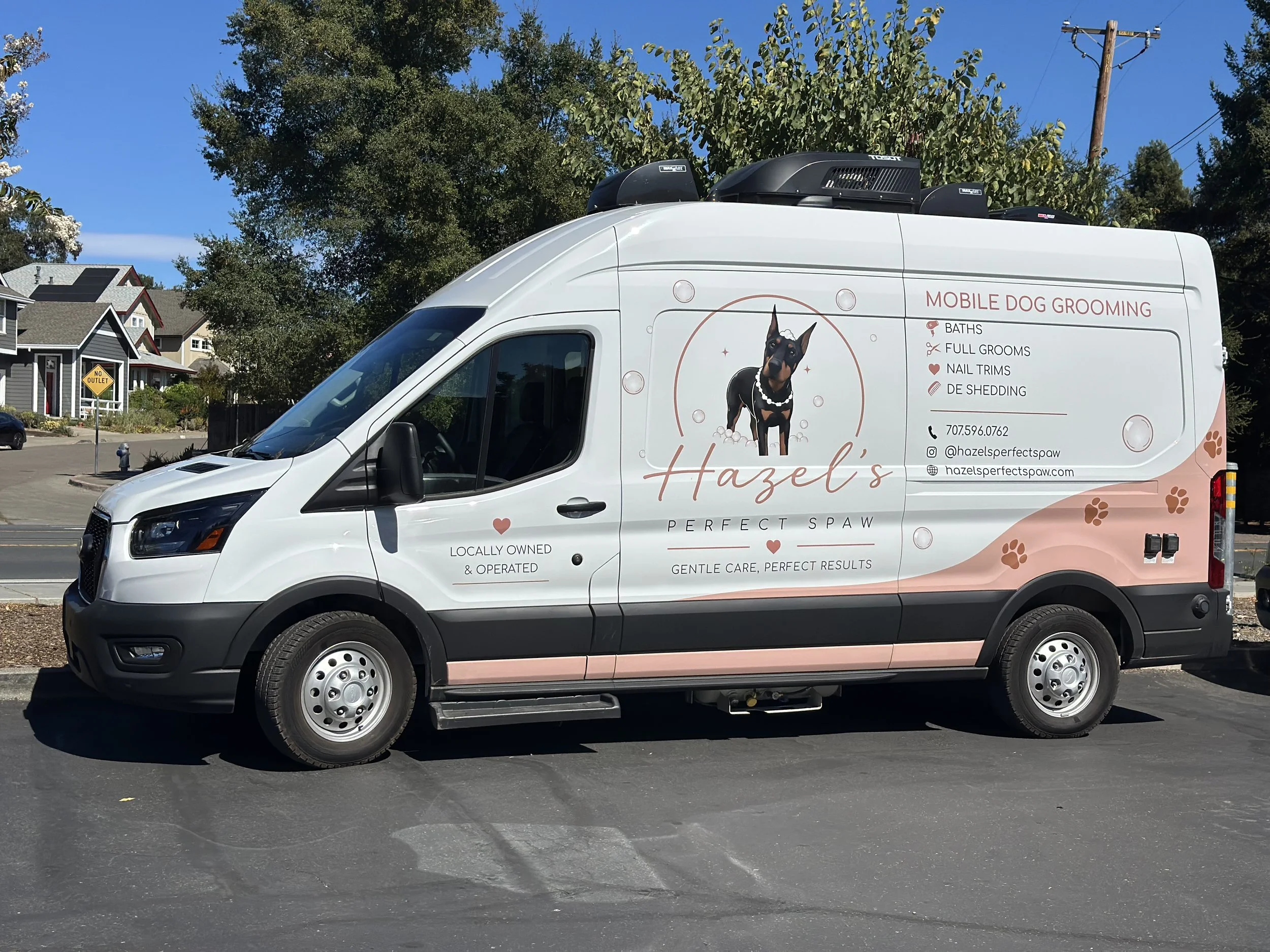 A white mobile dog grooming van with branding for Hazel's Perfect Spa, featuring a picture of a Doberman dog and services listed such as baths, full grooms, nail trims, and de-shedding. The van is parked on a street with trees, houses, and a utility pole in the background.