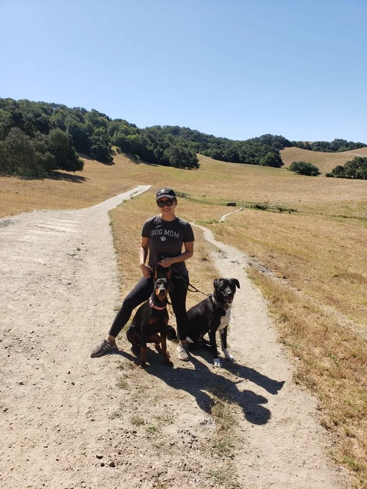 A woman holding two dogs on a dirt trail in a hilly, grassy area under a clear blue sky.