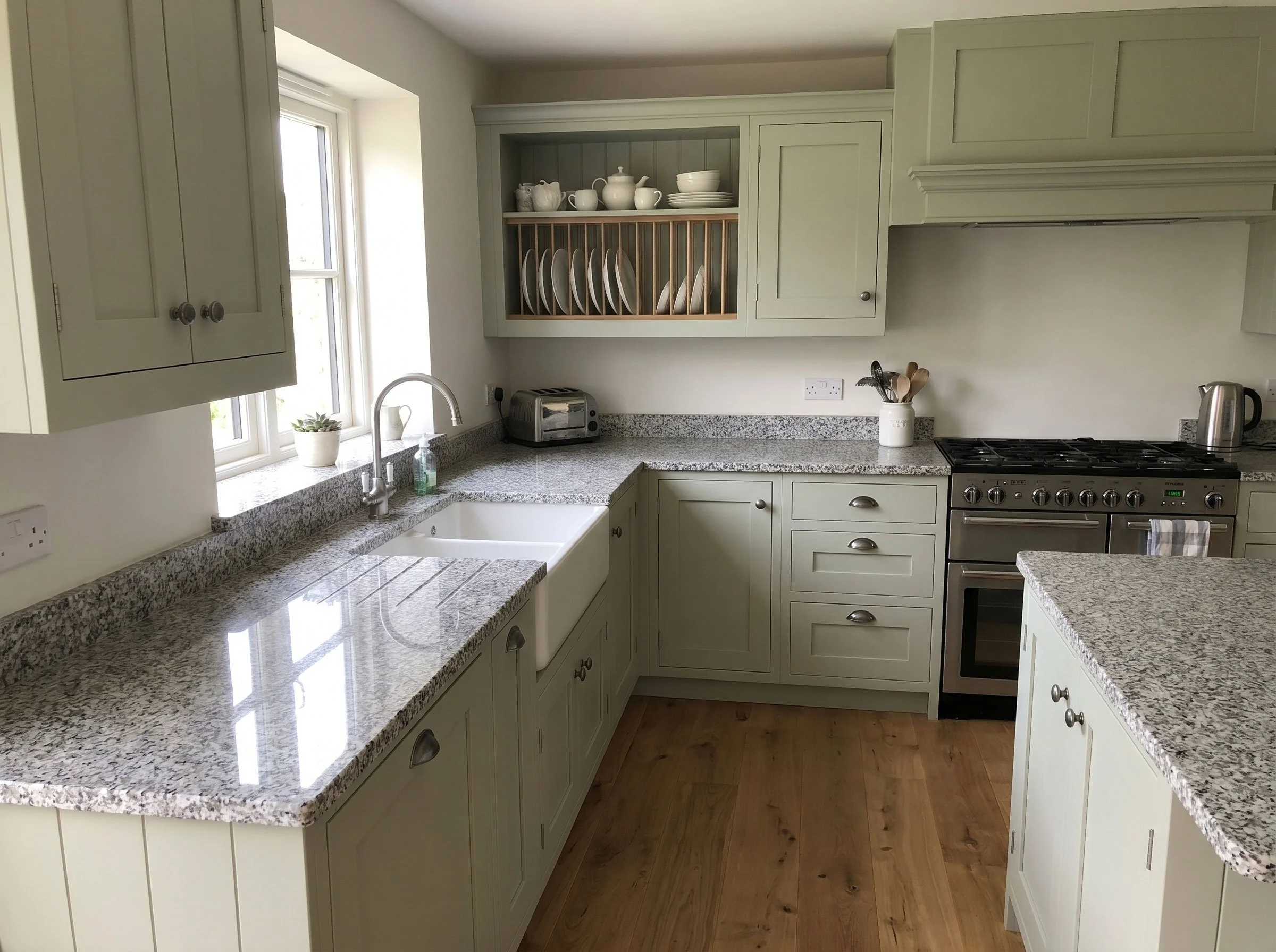A kitchen with granite countertops, cream-colored cabinets, a window above the sink, a toaster, a jar with cooking utensils, and a stove.