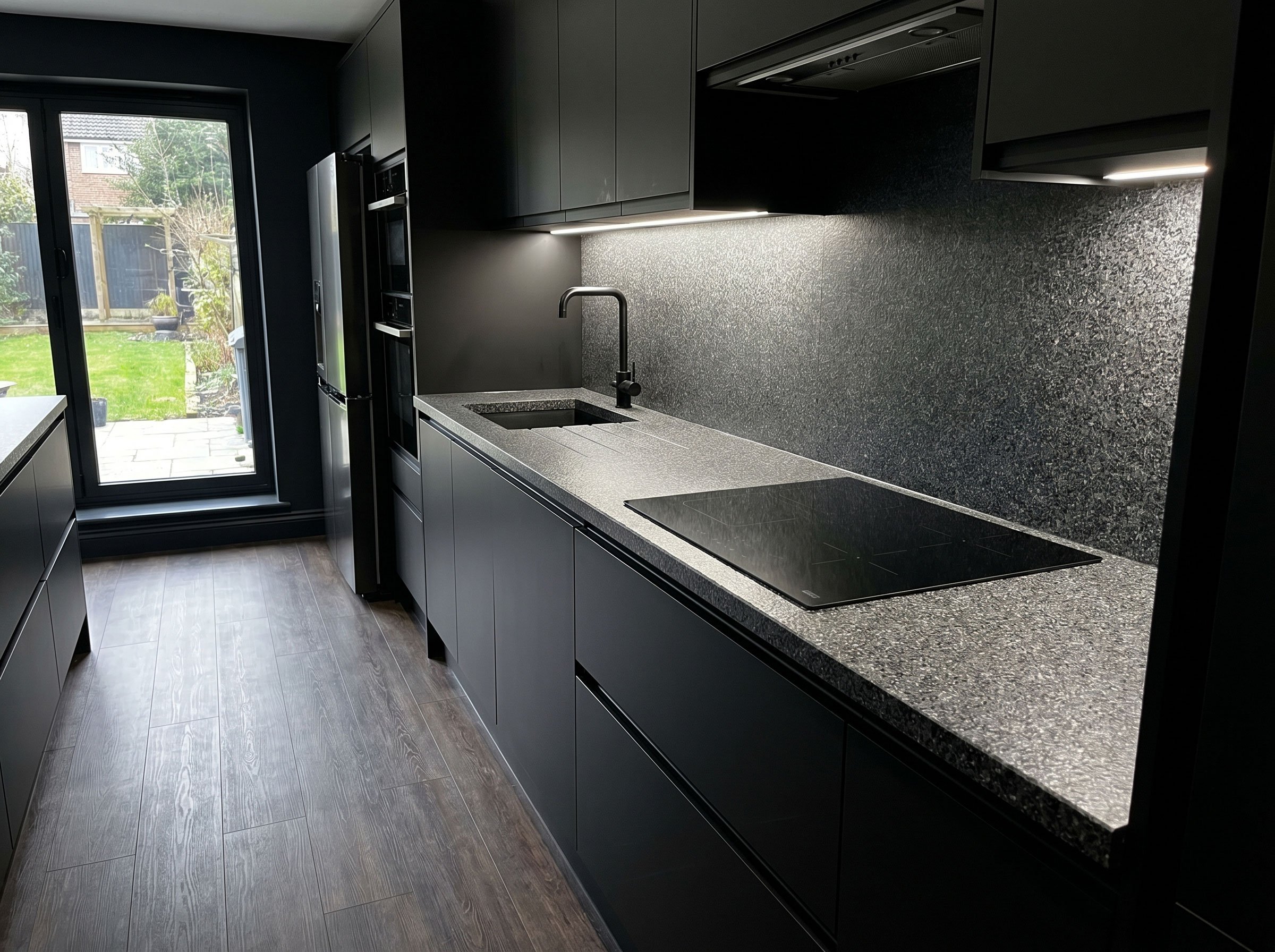 Modern kitchen with black cabinets and countertops, a sink with a black faucet, and a large glass door leading to a backyard with green grass and trees.