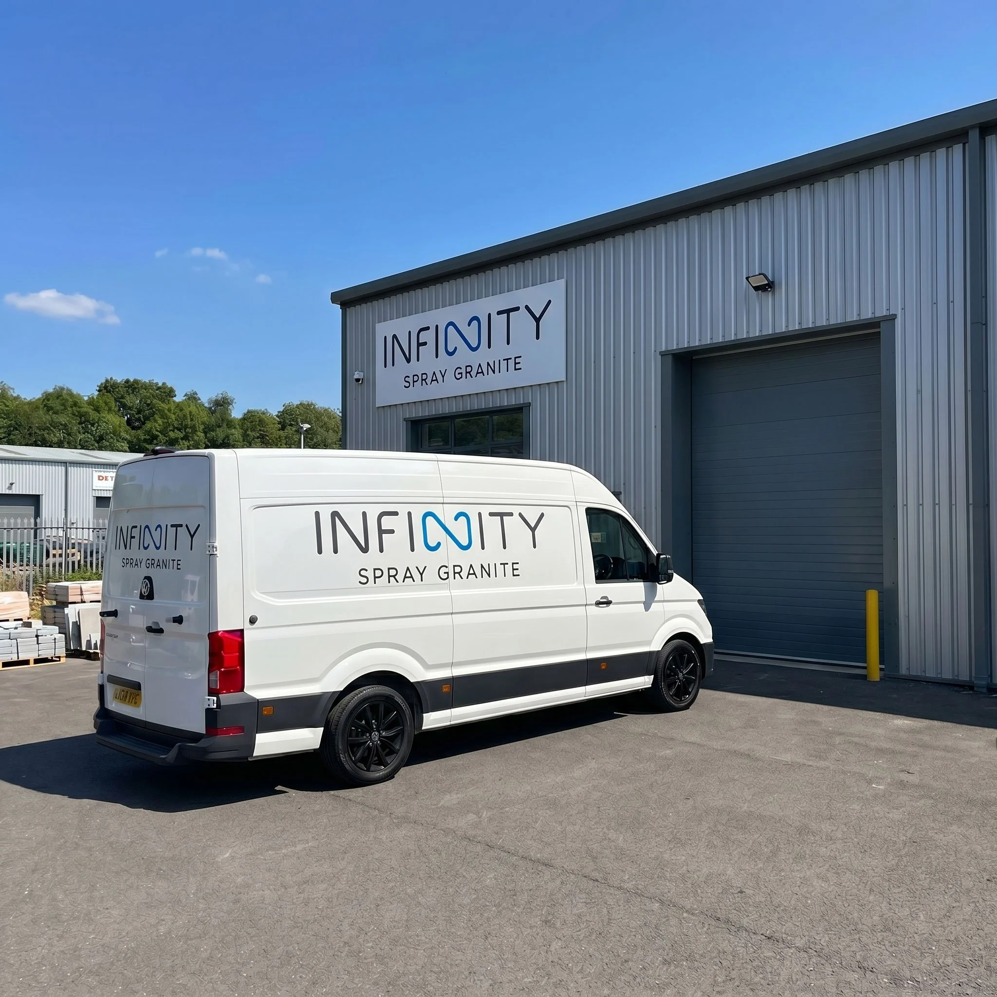 A white delivery van with 'INFINITY SPRAY GRANITE' logo parked in front of a grey industrial building with a sign for the same company, under a blue sky with a few clouds.