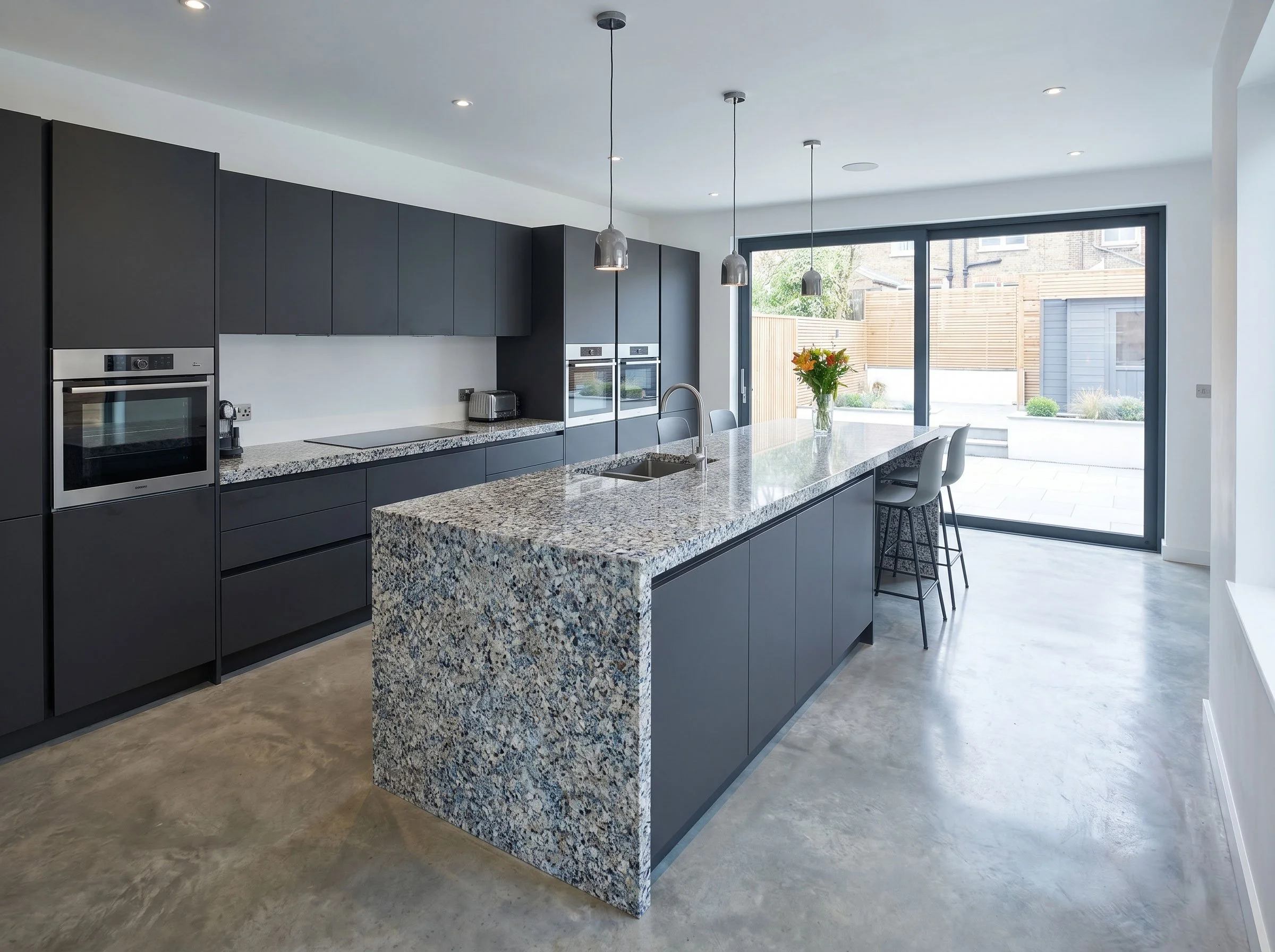 Modern kitchen with dark cabinets, granite island, stainless steel appliances, and large sliding glass door leading to backyard with wooden fencing.