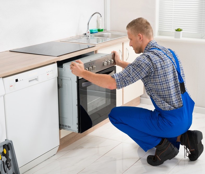 A man in blue overalls kneels in front of an oven, fixing or installing it in a kitchen.