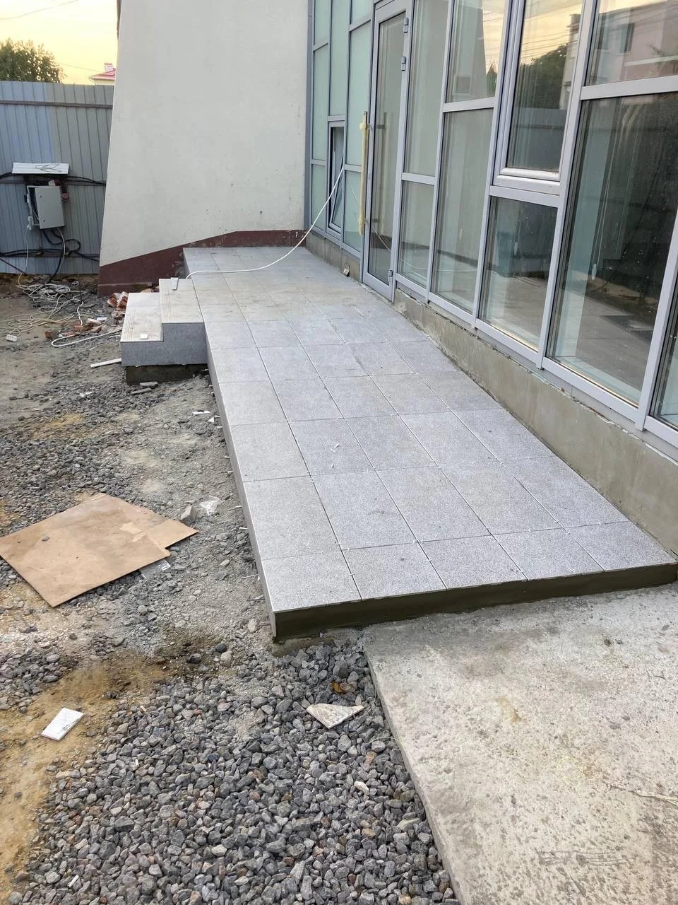 A newly tiled outdoor patio area with grey tiles in front of glass doors, surrounded by construction materials and uneven ground.