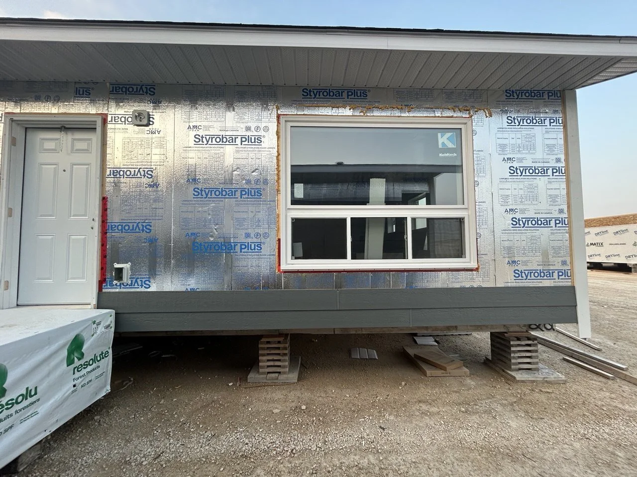 Construction site with a house under construction. The house is elevated on pallets, with a window and a door visible. Exterior walls are covered with insulation material labeled 'Styrobar Plus'.