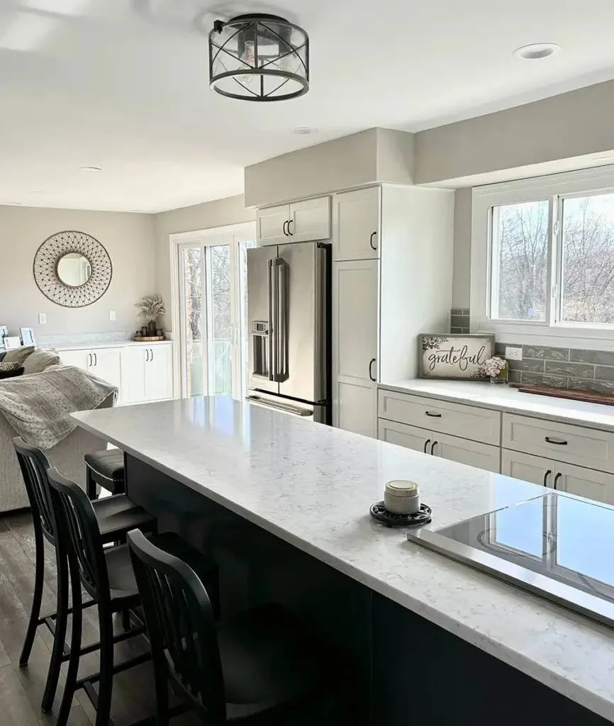 Modern kitchen with white cabinets, a marble countertop island with black chairs, stainless steel refrigerator, and windows showing a winter outdoor scene.