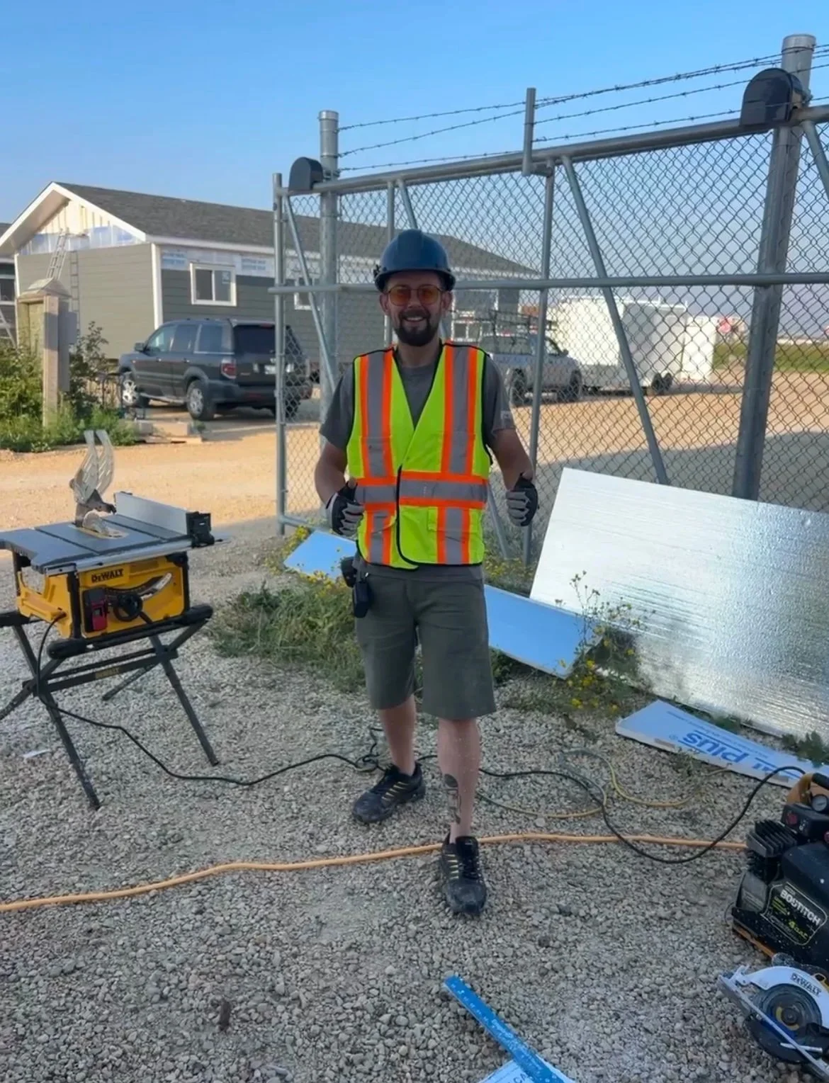 A man wearing a yellow safety vest, helmet, sunglasses, and gloves standing on a construction site with tools and metal sheets around him.