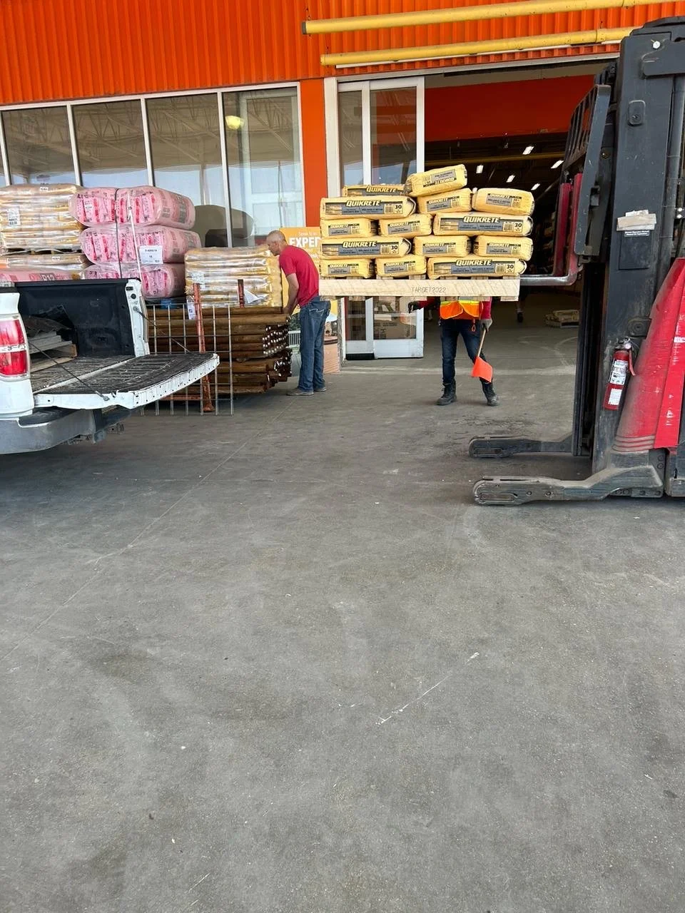 People unloading bags and rolls of insulation or construction material from a forklift outside a store.