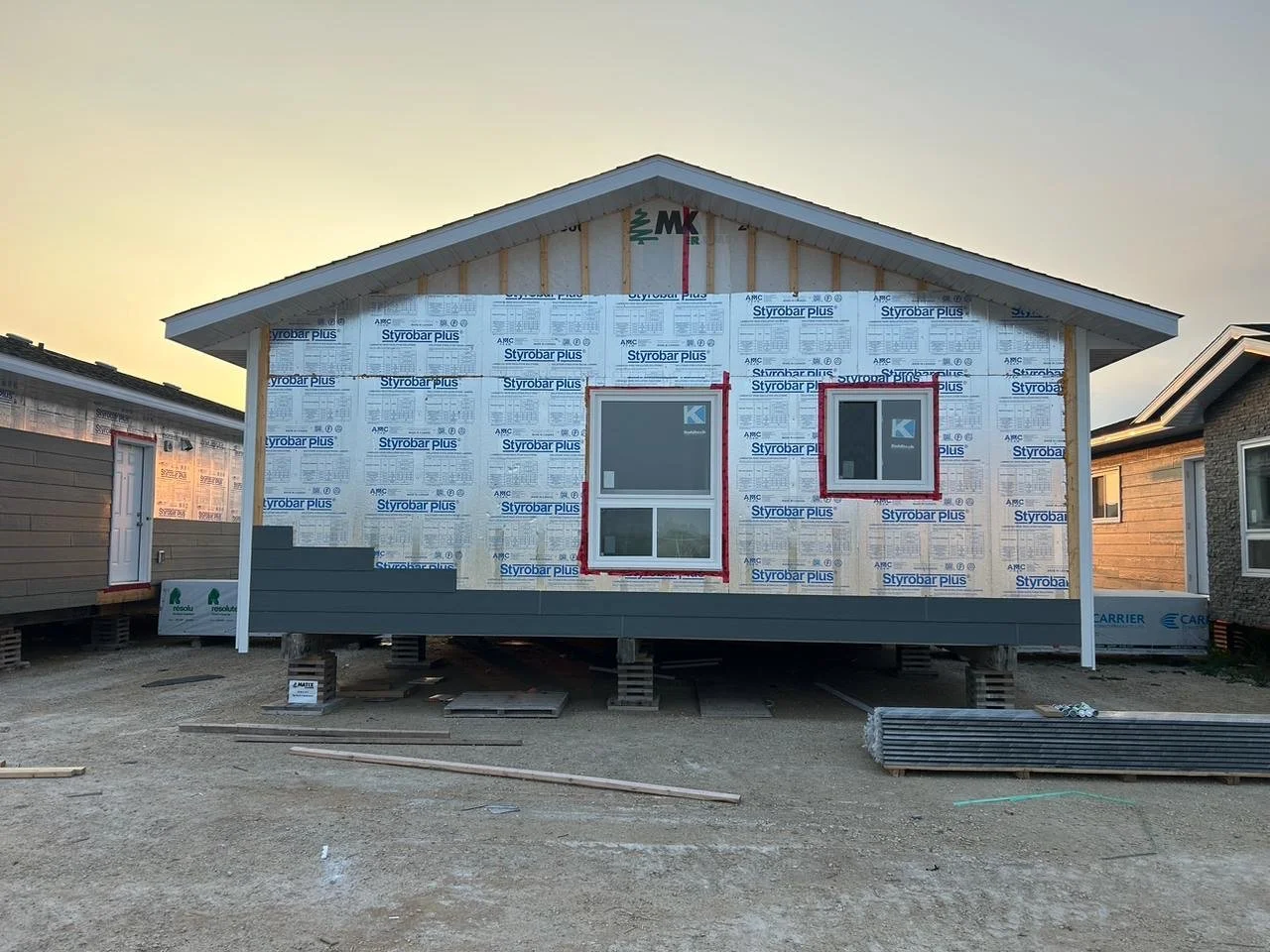 Front view of a house under construction with exterior sheathing and two windows installed, on a dirt lot with construction materials in the foreground.