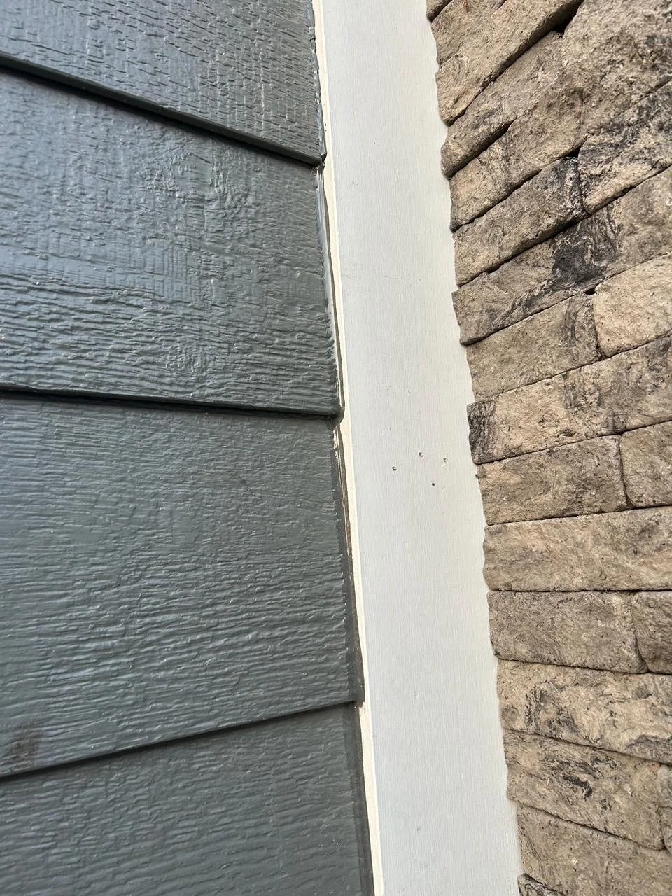 Close-up view of the intersection of different exterior wall materials: gray textured siding on the left, smooth white trim in the middle, and beige stacked stone on the right.