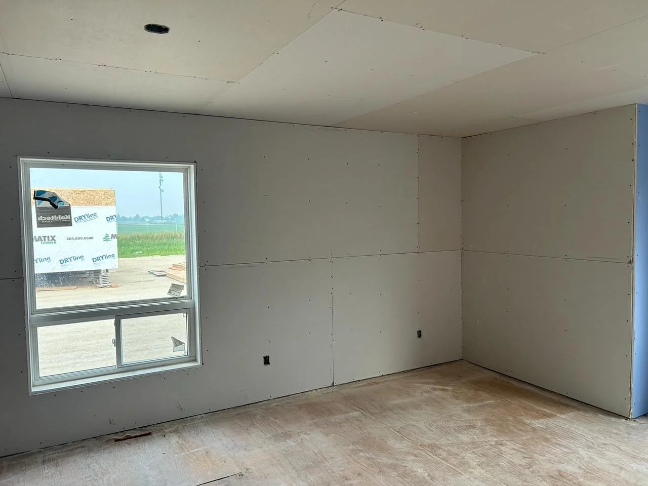 Interior view of a room under construction with drywall panels installed on the walls and ceiling and plywood flooring, with a window showing an outdoor construction site.