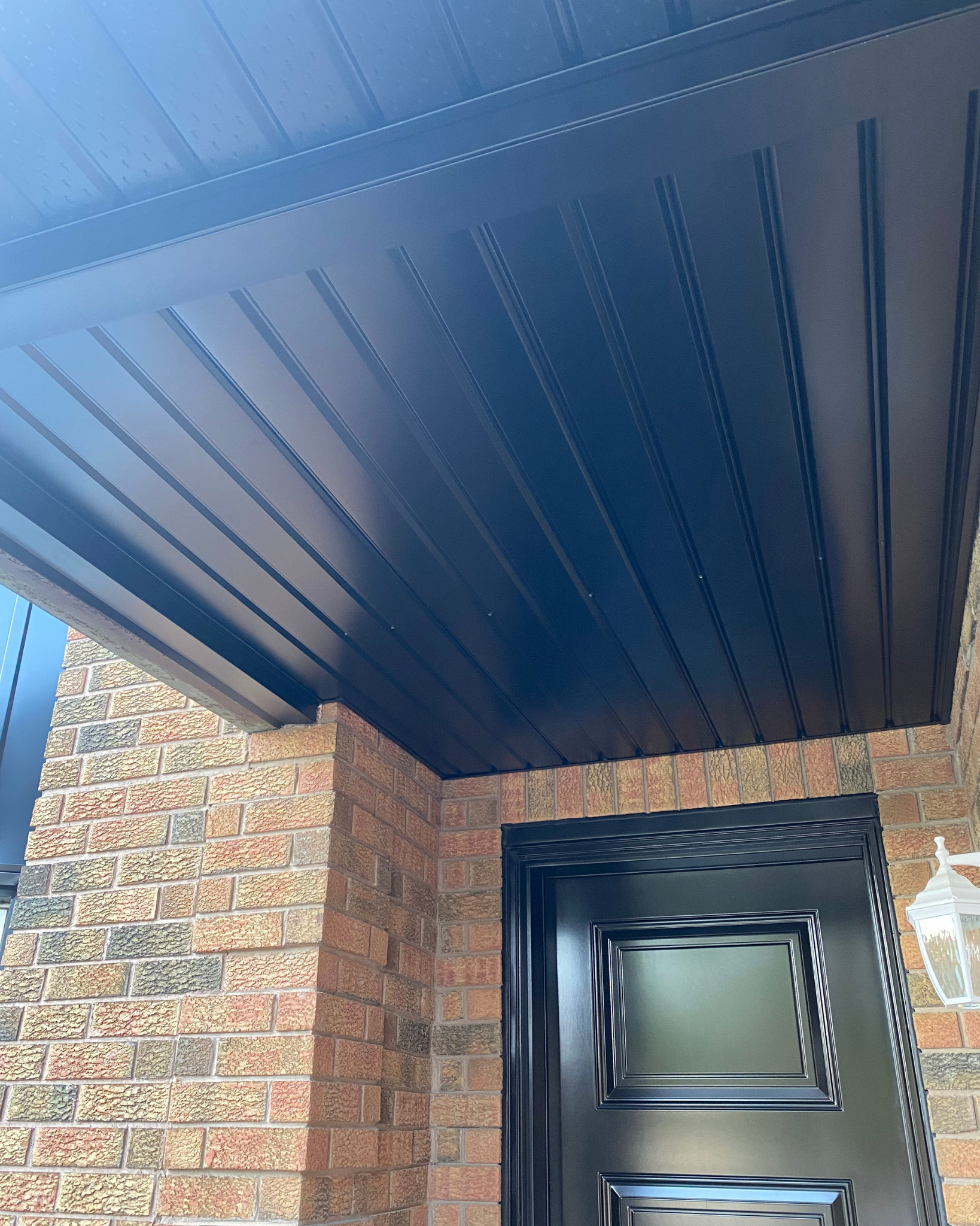 Photo of a brick house with a black door and black framing, featuring an outdoor brick column and a blue metal awning.