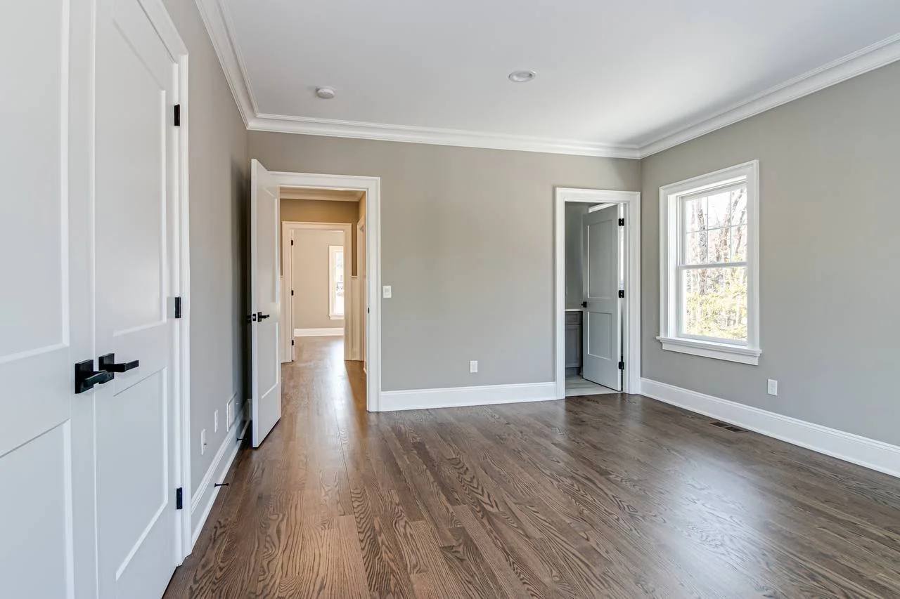Empty room with hardwood floor, gray walls, white trim, and multiple doorways, one leading to another room with similar decor, and a window allowing natural light.