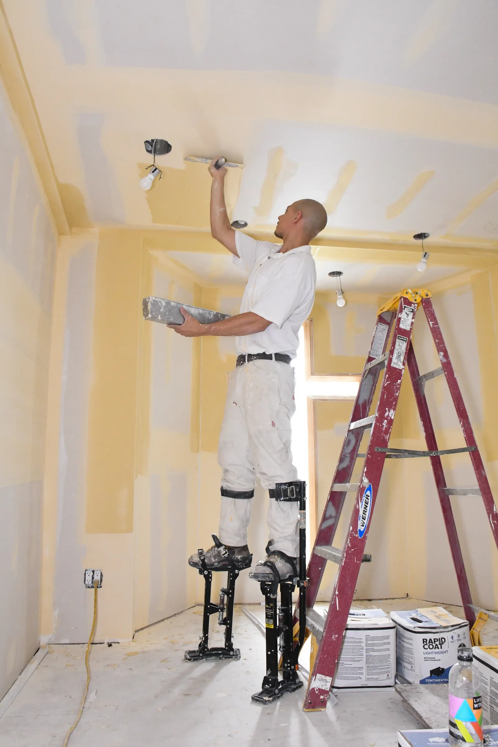 Man standing on a scaffolding while painting a ceiling in a room. The room has yellow painted walls and there are construction supplies on the floor.