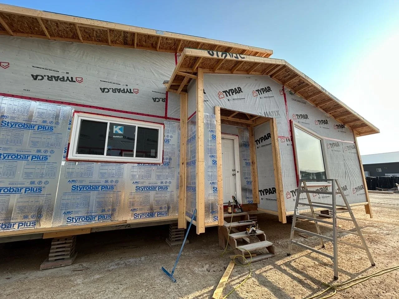 Under construction tiny house with wooden stairs, ladder, and window installed, wrapped in construction insulation and weatherproofing on a dirt lot.
