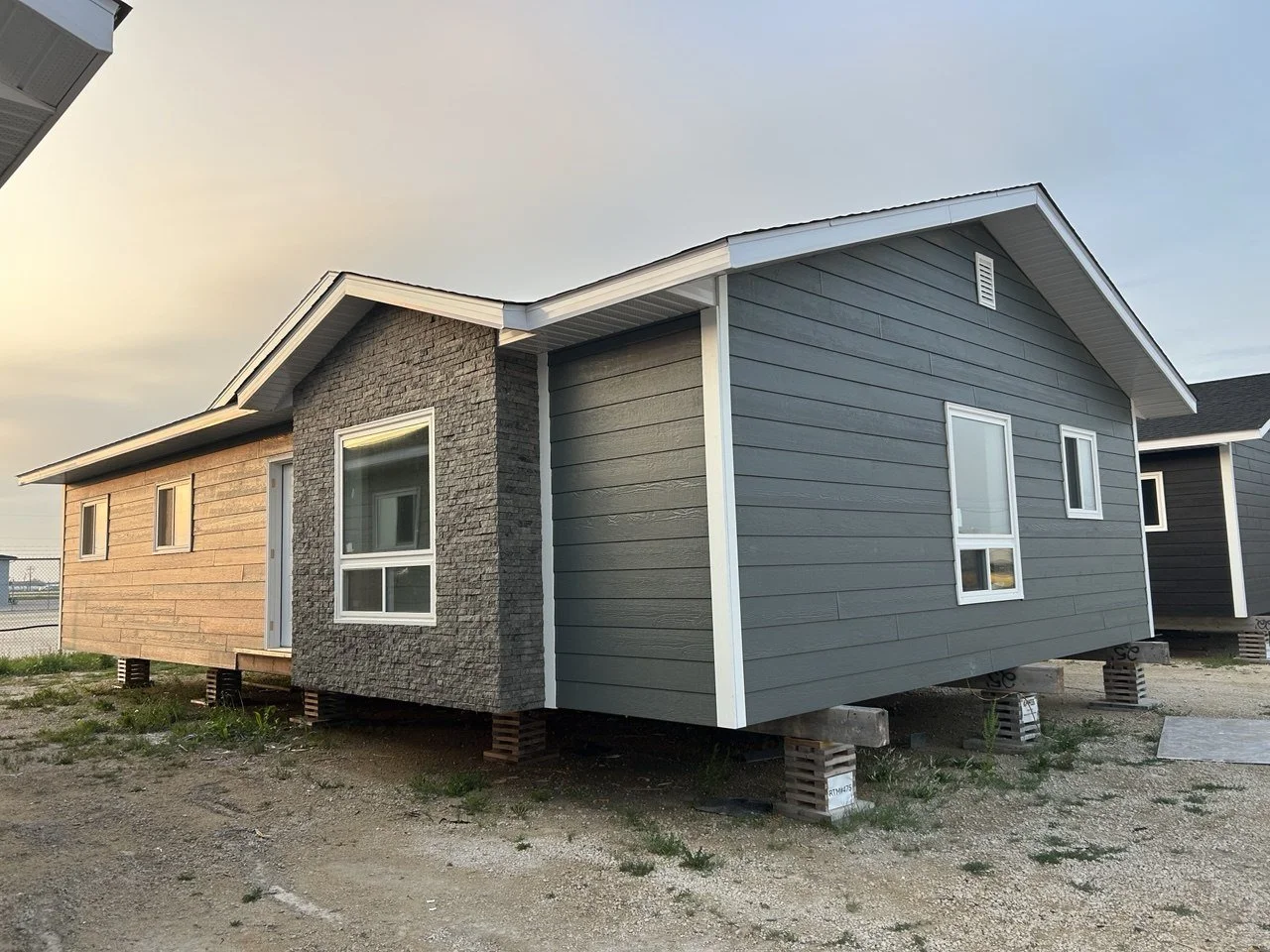 A house under construction with gray horizontal siding, stone accents, and white window frames, elevated on concrete blocks on a gravel lot.