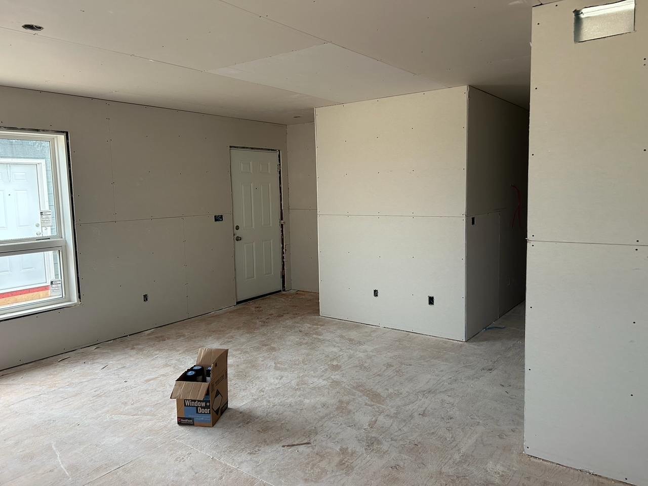 Interior of a room under construction with unfinished drywall, a window, a door, electrical outlets, and a cardboard box on the floor.