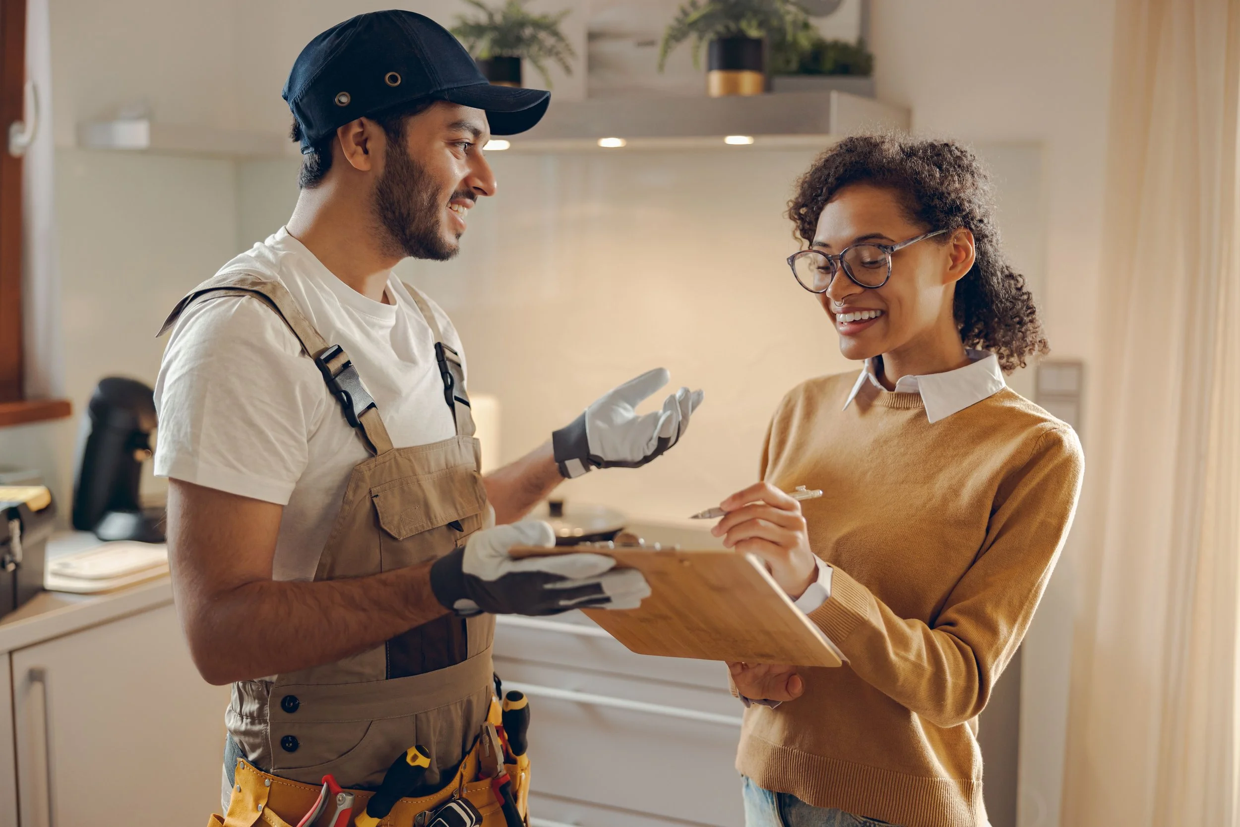 A man and a woman are indoors, engaging in a friendly conversation. The man, dressed in a work uniform with a tool belt, is showing a tablet to the woman who is smiling and holding a clipboard. The setting appears to be a home or kitchen environment.