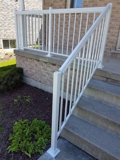 White staircase railing with vertical bars on an outdoor steps leading to a porch, with brick exterior wall and green shrubbery nearby.