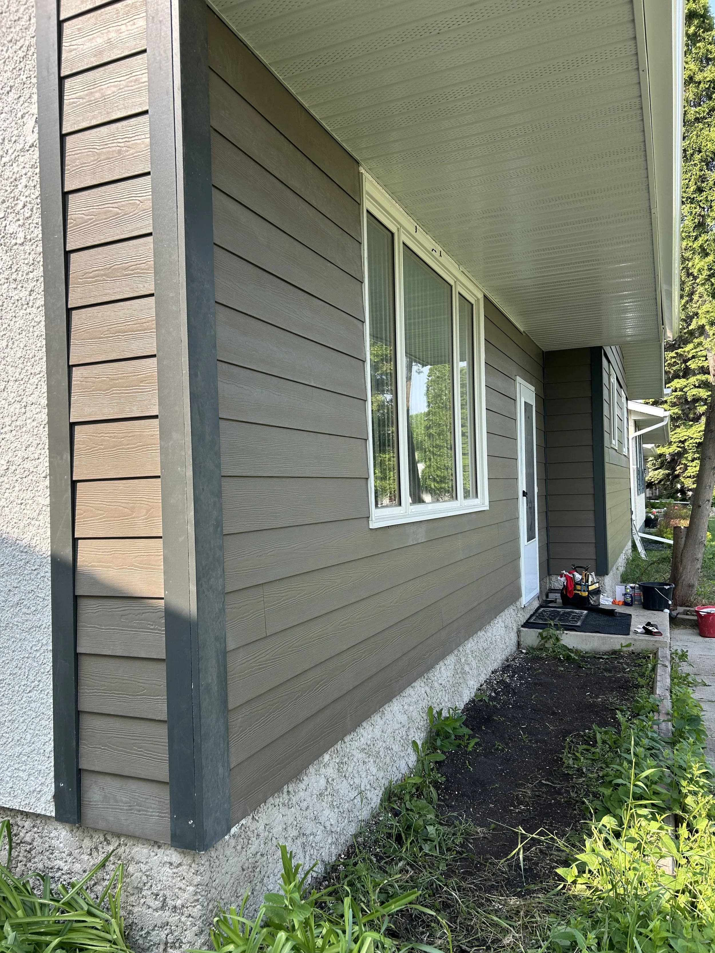 Front view of a house with beige horizontal vinyl siding, white-framed windows, and a small front yard with gardening tools and plants.