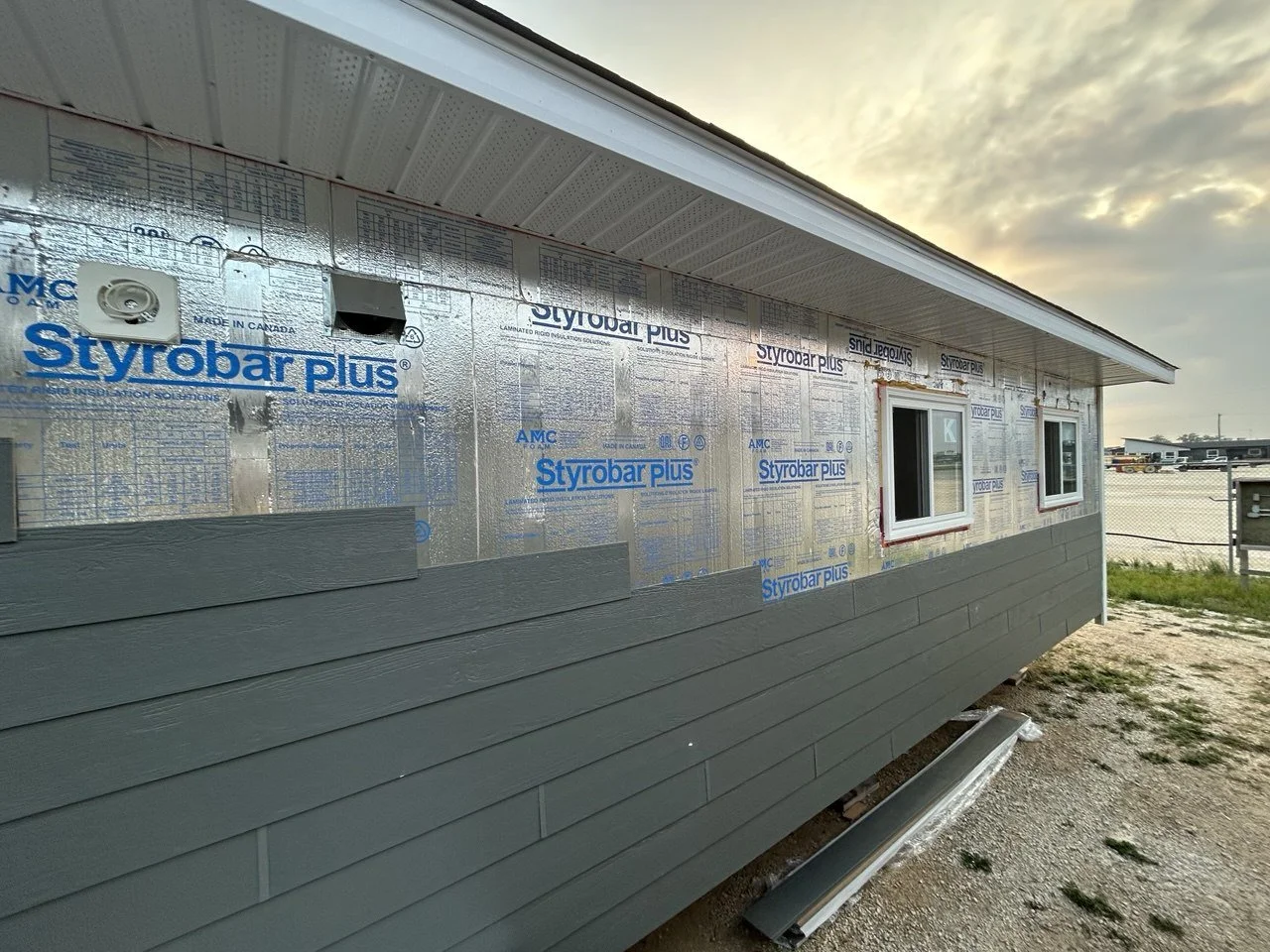 Side of a house under construction with vinyl siding partially installed, two small windows, and insulation visible.