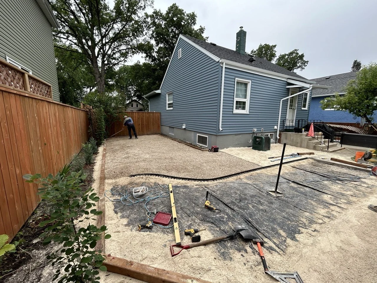 Backyard under construction with a worker leveling gravel, blue house with white trim, wooden fence, and construction tools and materials on the ground.