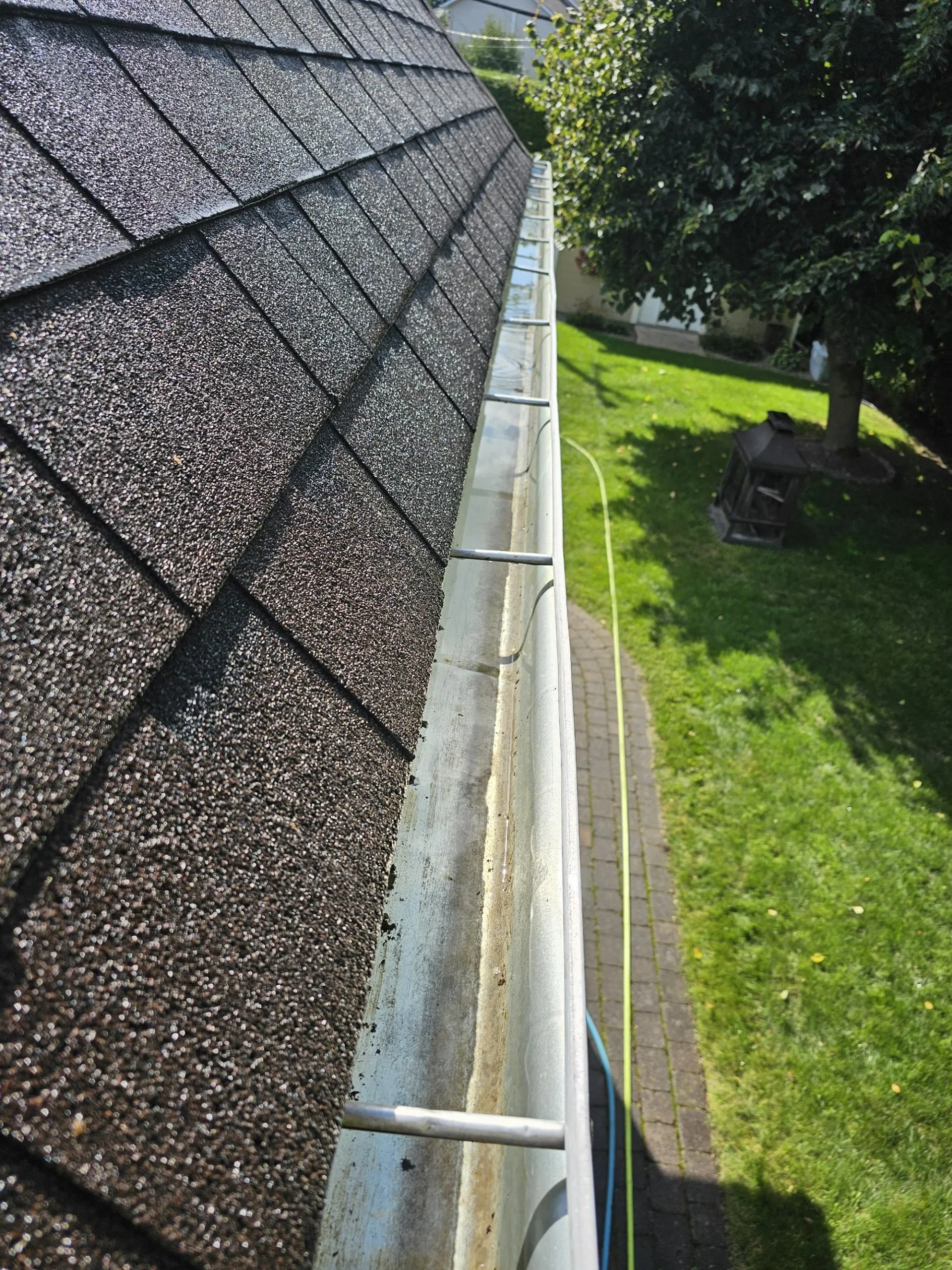 Close-up of a section of a house roof with asphalt shingles, viewed from the gutter area. The background shows some trees, a walkway, and a grassy lawn.
