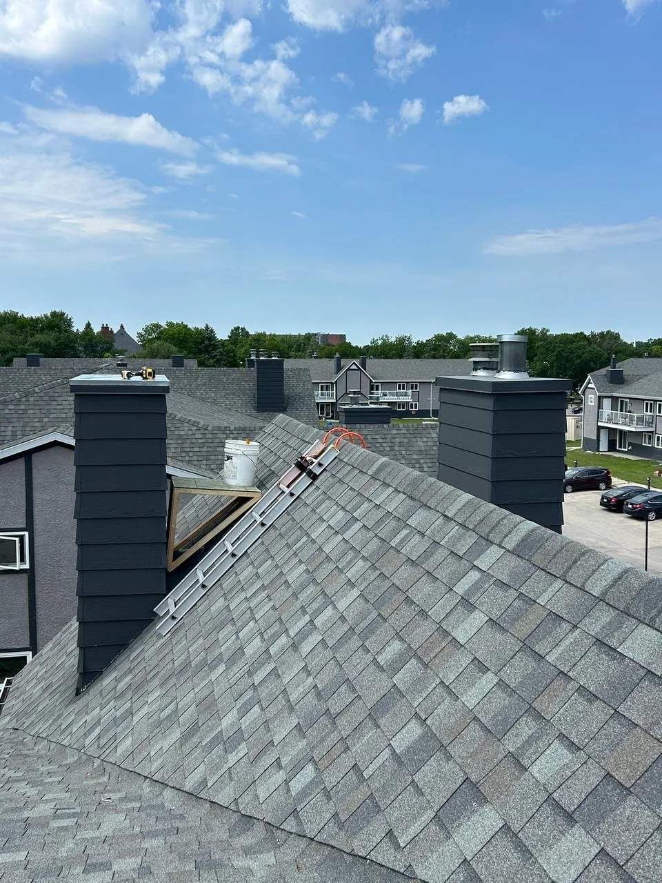 View of a rooftop with gray shingles, two black chimneys, a ladder, and a skylight with a wooden frame. In the background, there are more buildings, trees, and a parking lot.