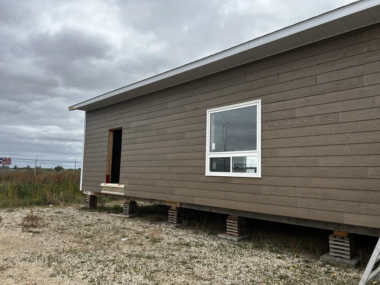 A house under construction with brown horizontal siding, an open doorway without a door, a single window with a white frame, and concrete blocks supporting the structure. The ground is gravel and dirt, with some grass and weeds. Overcast sky with gra