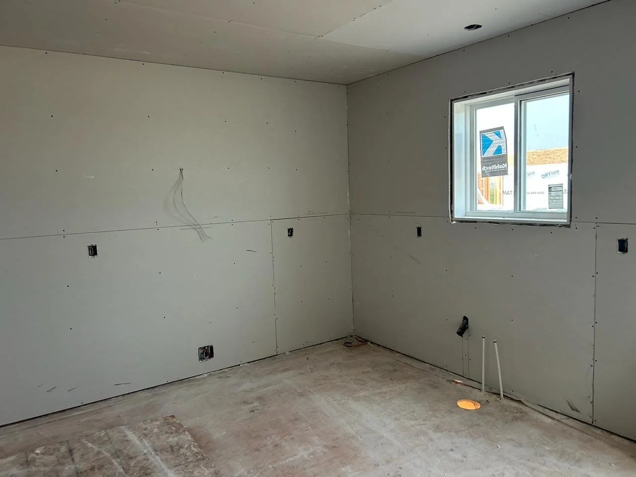 Room under construction with drywall installed, electrical wiring visible, and a window showing an outside view of a building with a sign that reads 'DryLine.'