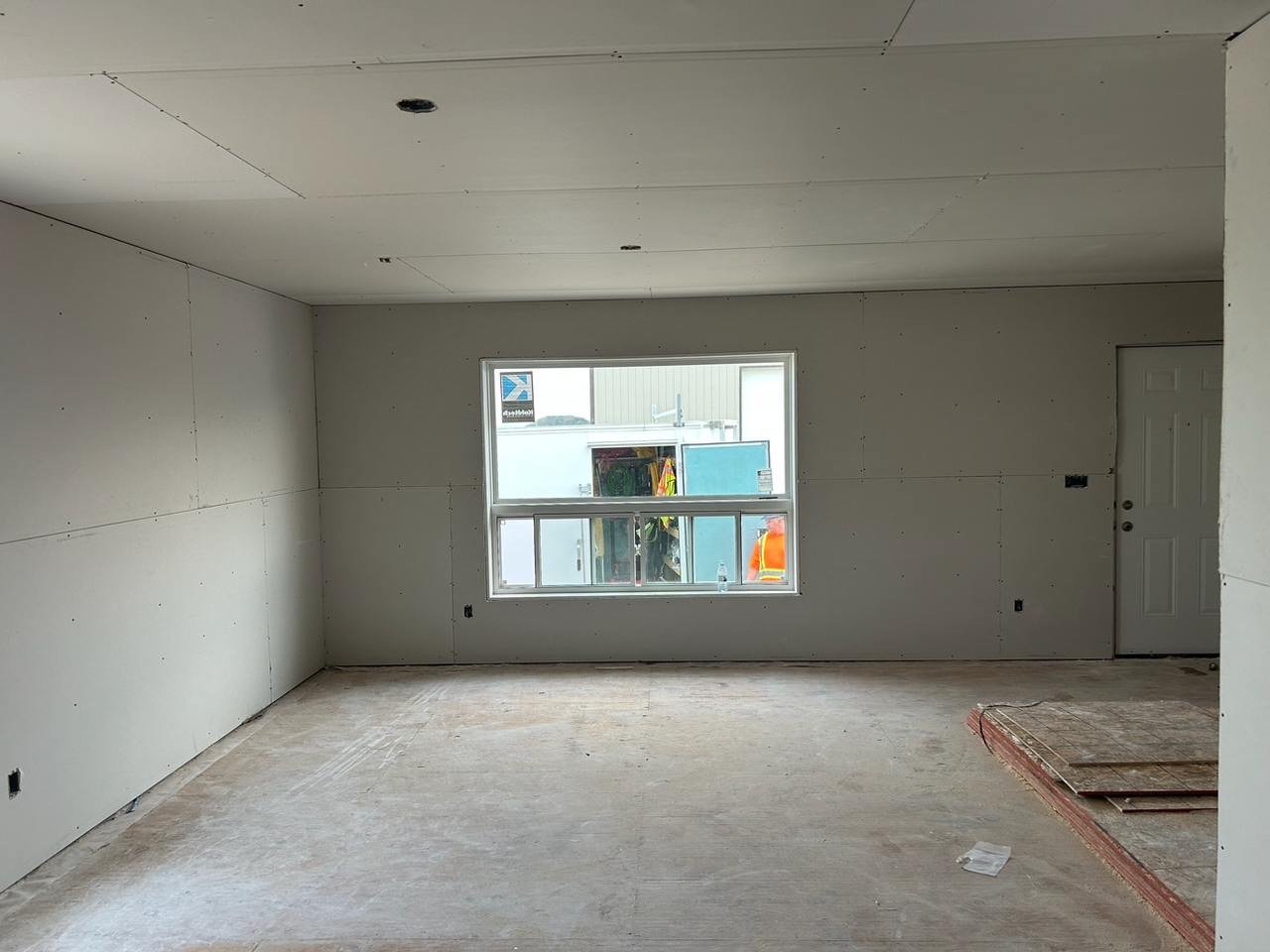 Room under construction with drywall installed, window in the center, and door on the right, construction materials on the floor.