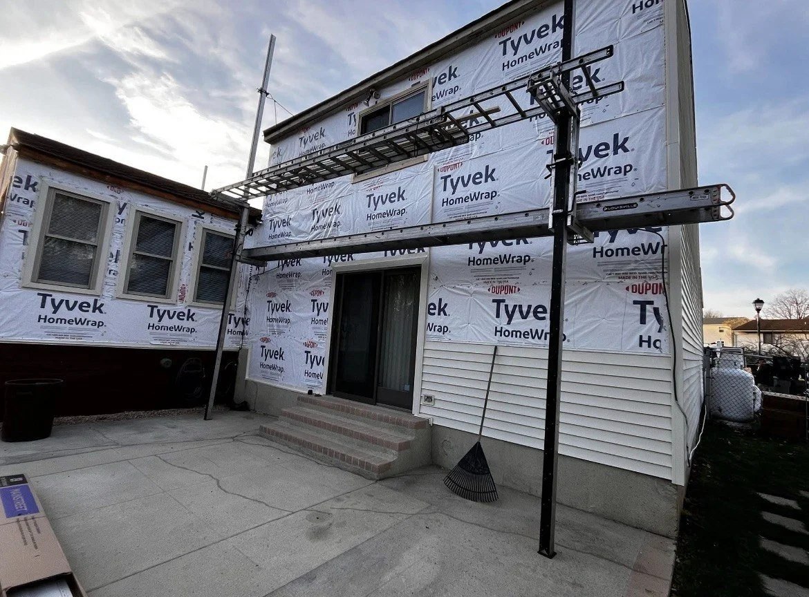 House under construction with building wrap labeled 'Tyvek HomeWrap'. There are steps leading to a sliding door, and metal scaffolding or support structure above the entrance. A rake leans against the house, and an antenna is attached to the house. The sky is partly cloudy.
