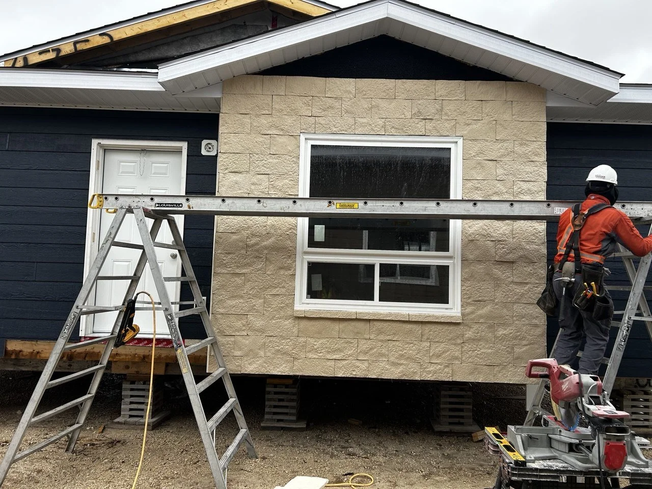 Construction worker on a ladder installing or repairing exterior siding on a house with a large window. The house has a two-tone facade with beige stone and dark blue siding, and the worker is wearing safety gear and a white helmet.