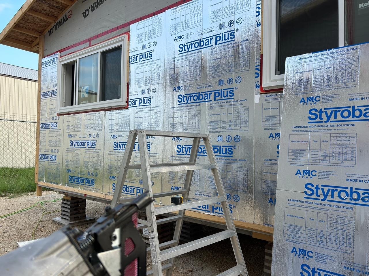 Construction site showing a house with insulation panels marked Styrobar Plus and AMC Foam, a window installed, a ladder, and a power tool in the foreground.
